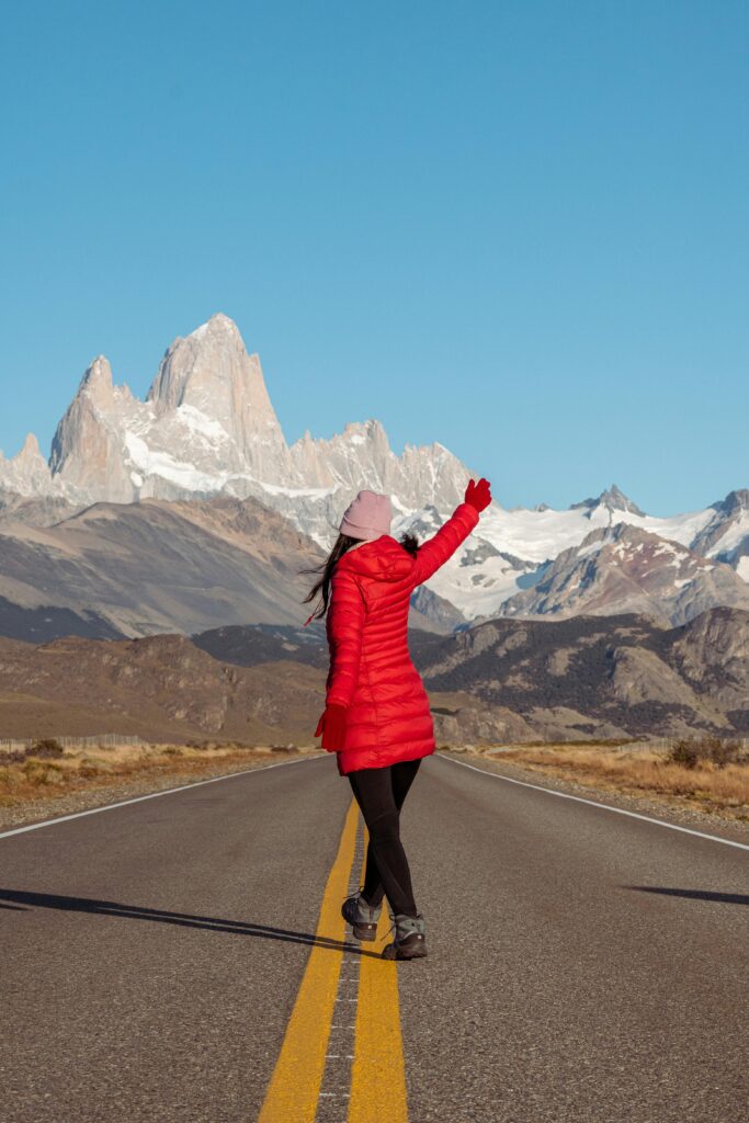 A woman in a red jacket stands on a road leading to towering mountains, embracing adventure.